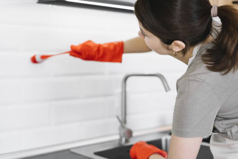 A woman is cleaning oil stains from the kitchen wall with a brush